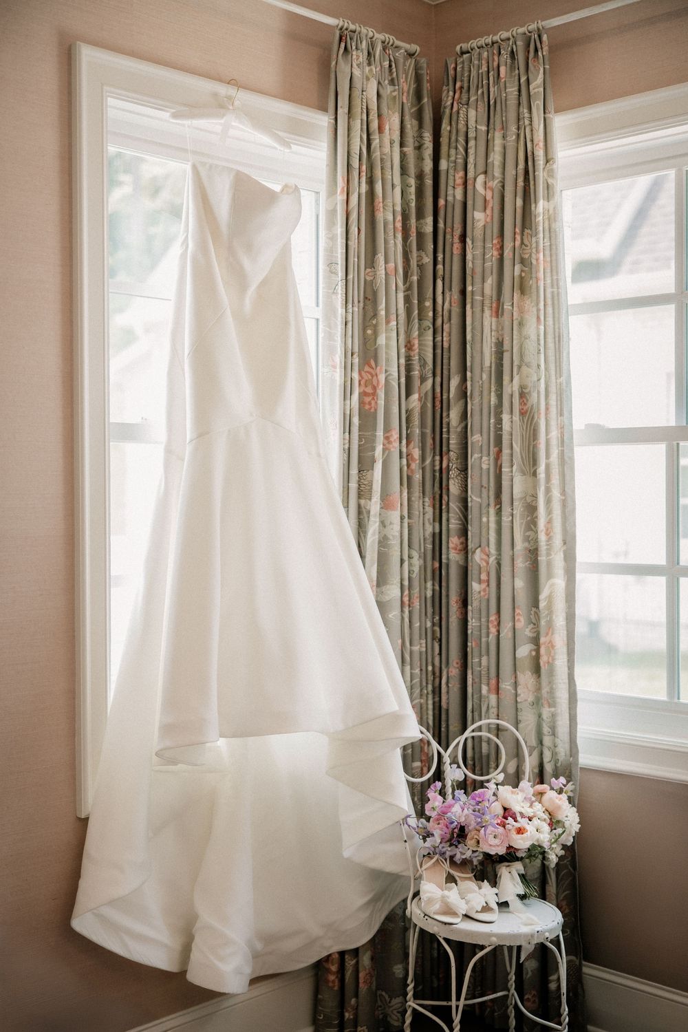 White wedding dress hanging by window with floral curtains in natural light.