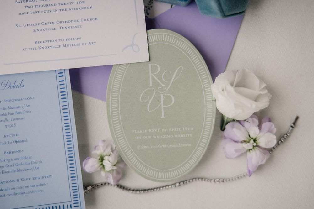Wedding invitation suite with sage green details and delicate white floral accents arranged on a purple background.