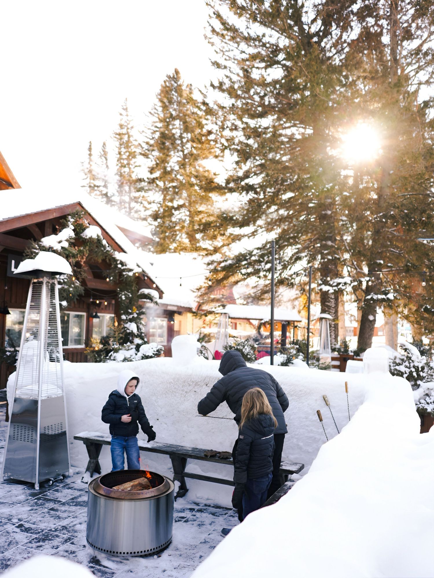 People gather around outdoor hot tubs surrounded by snowy landscape and pine trees at sunset in winter.