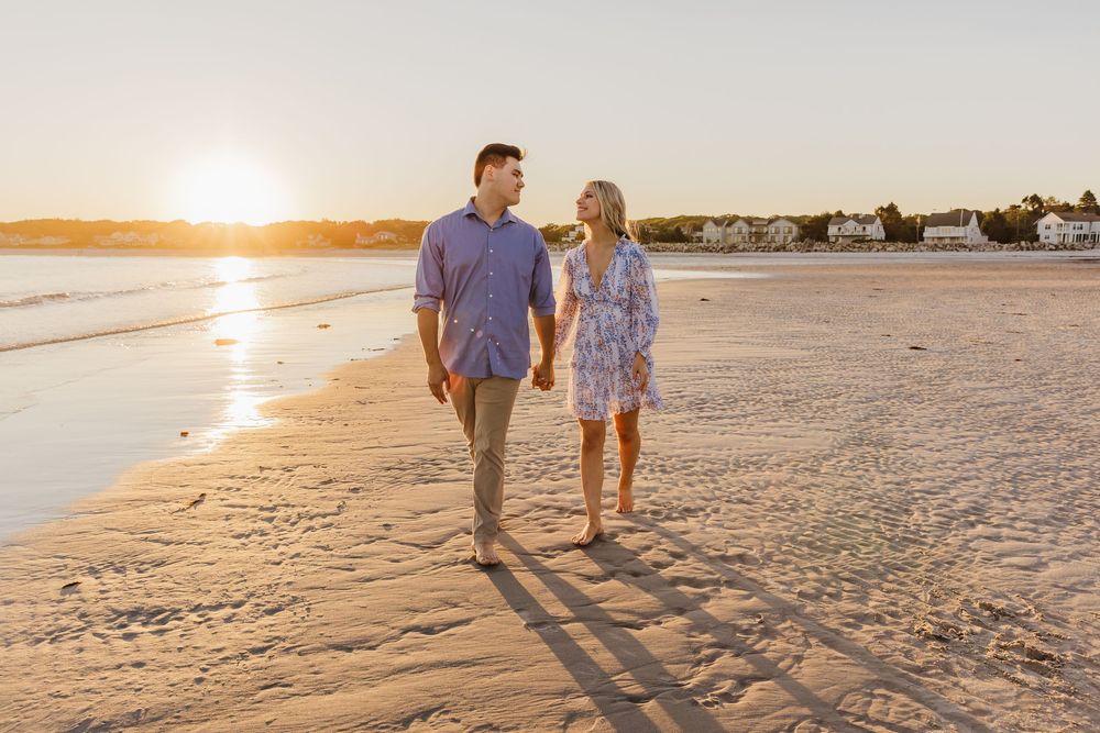 Couple walking on a sandy beach at sunset casting long shadows on the shore as the sun glows on the horizon.