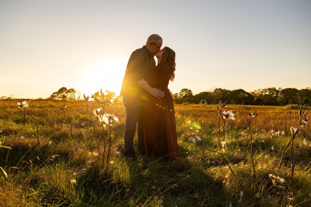 Silhouettes of two people sitting in a golden field at sunset while sun rays filter through tall grass.