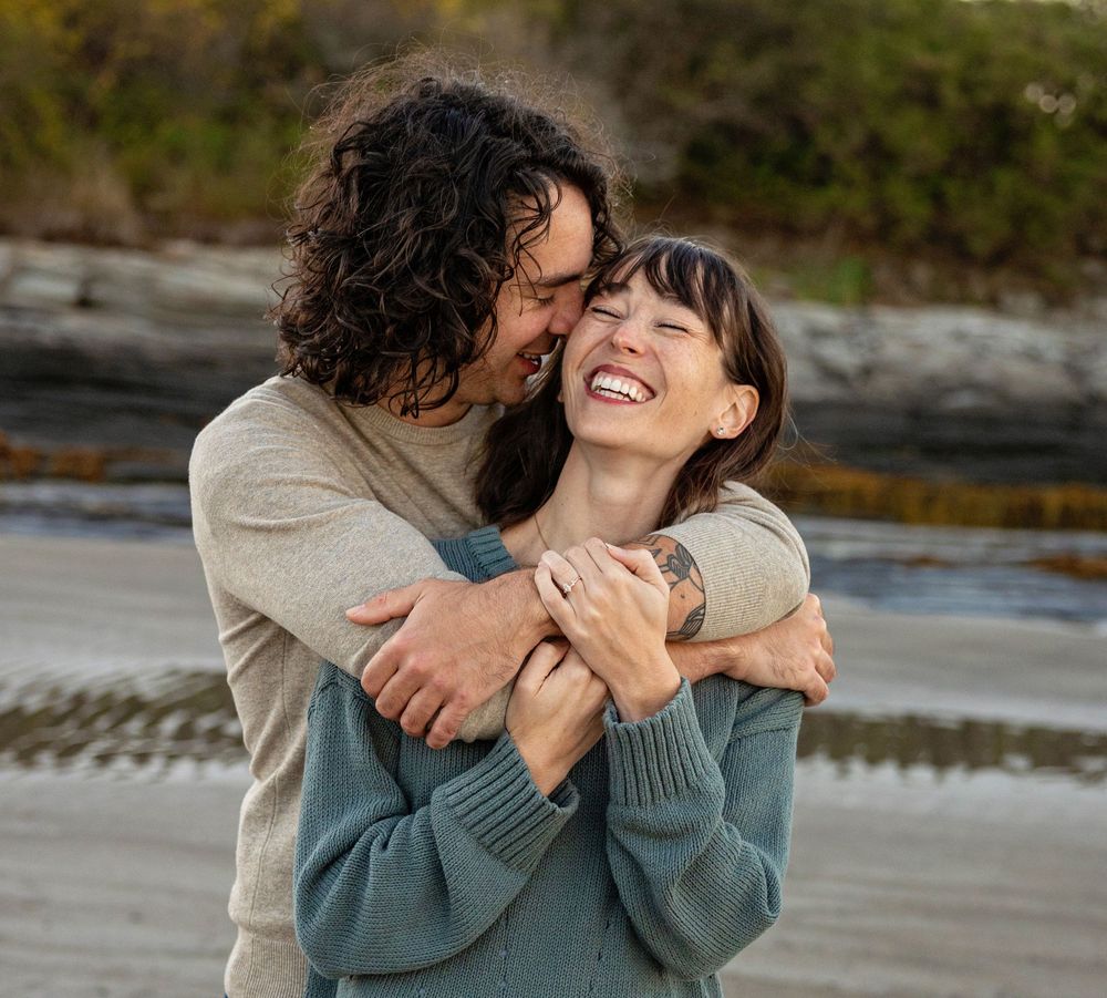 A romantic couple embraces and laughs together on a rocky beach during an outdoor photo session.