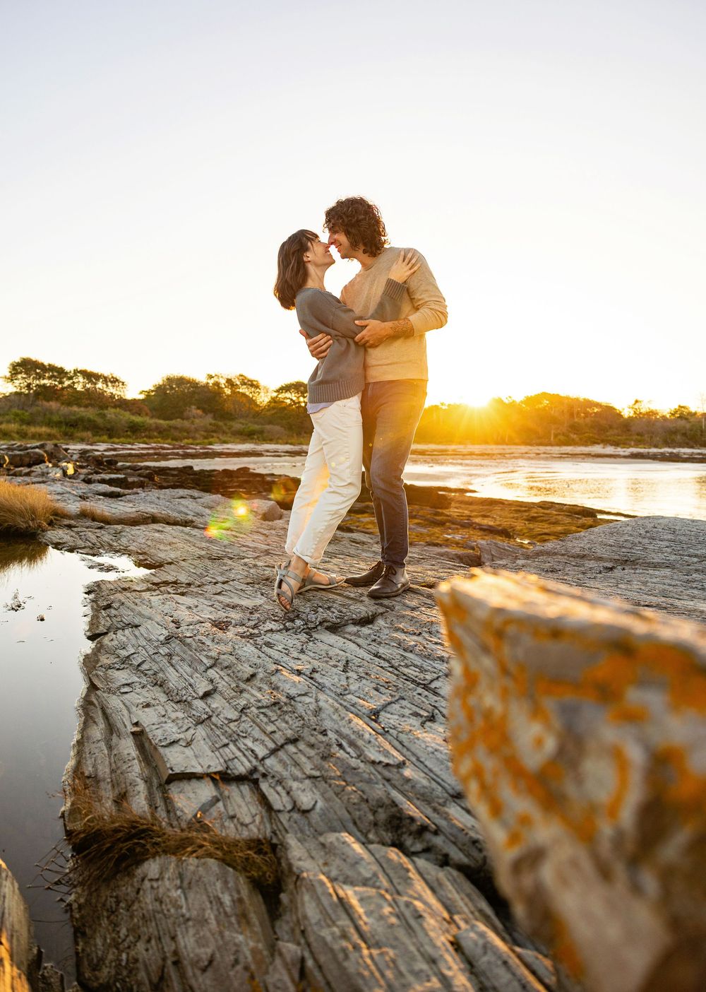 Couple embraces on rocky shoreline at sunset with golden light reflecting off coastal waters.
