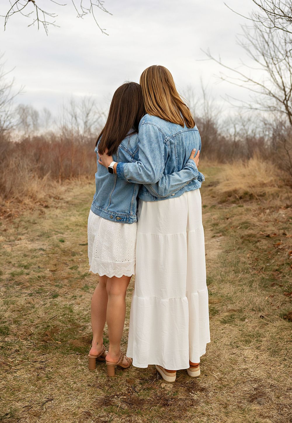 Two people in white dresses and denim jacket embrace while standing in a brown autumn field on an overcast day.