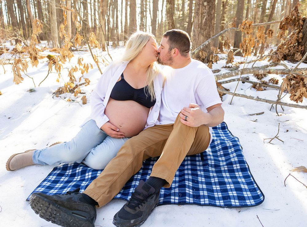 A panoramic winter scene shows a couple sitting together on a blue checkered blanket in the snowy woods.