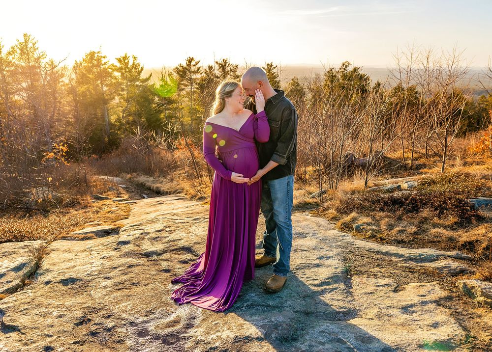 A couple in purple and blue attire embrace during golden hour on a rocky mountain overlook at sunset.