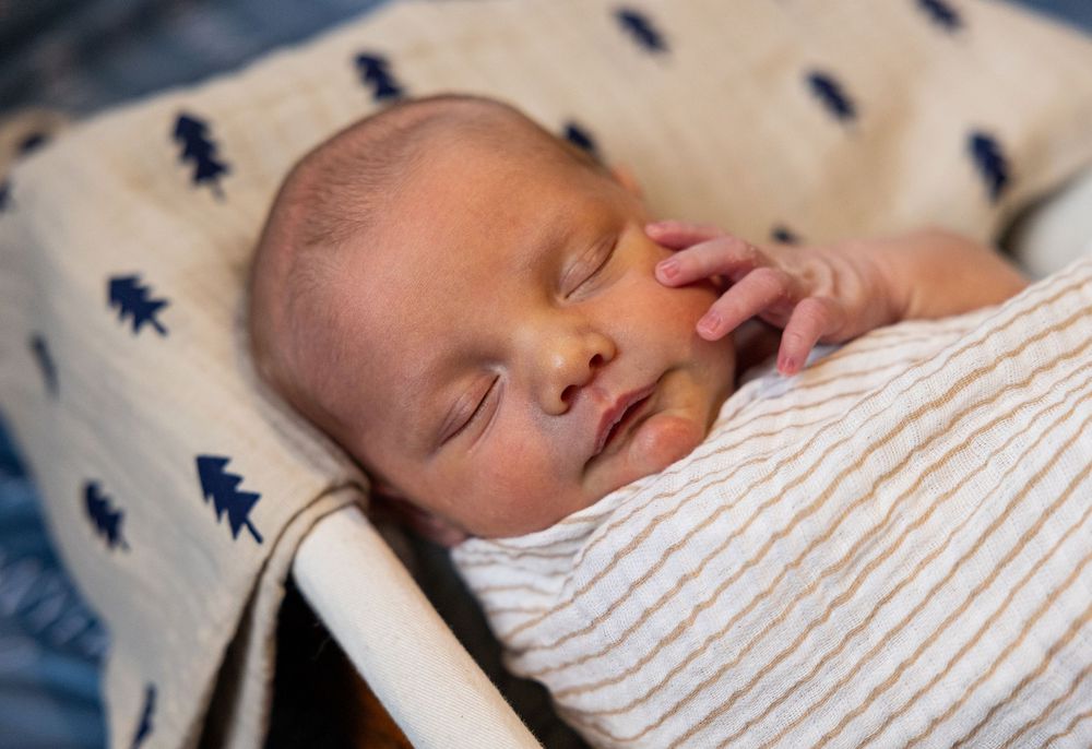 Sleeping baby wearing a striped onesie rests peacefully on navy blue patterned bedding.