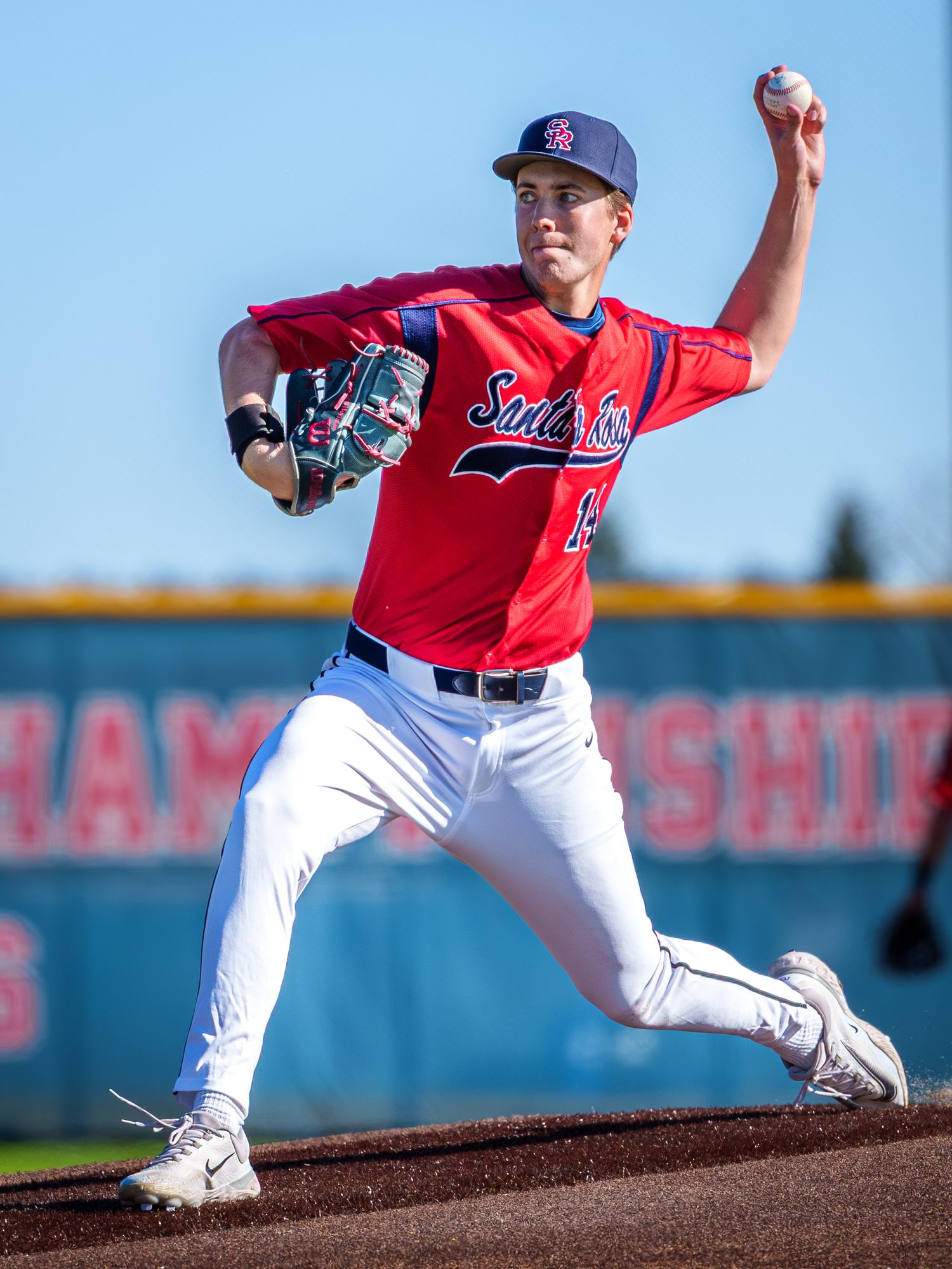 Baseball pitcher in red jersey and navy cap winds up to throw from the mound during a game against blue sky backdrop.
