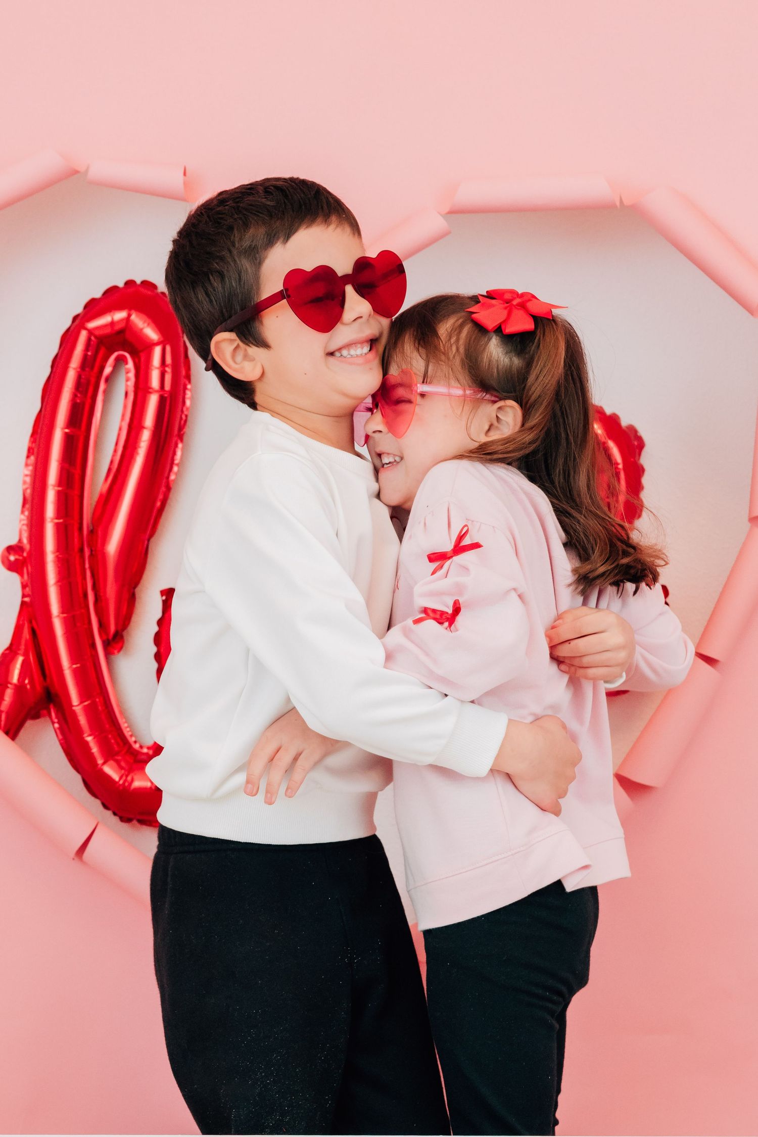 Two children wearing red heart sunglasses hug and laugh in front of a pink heart backdrop.