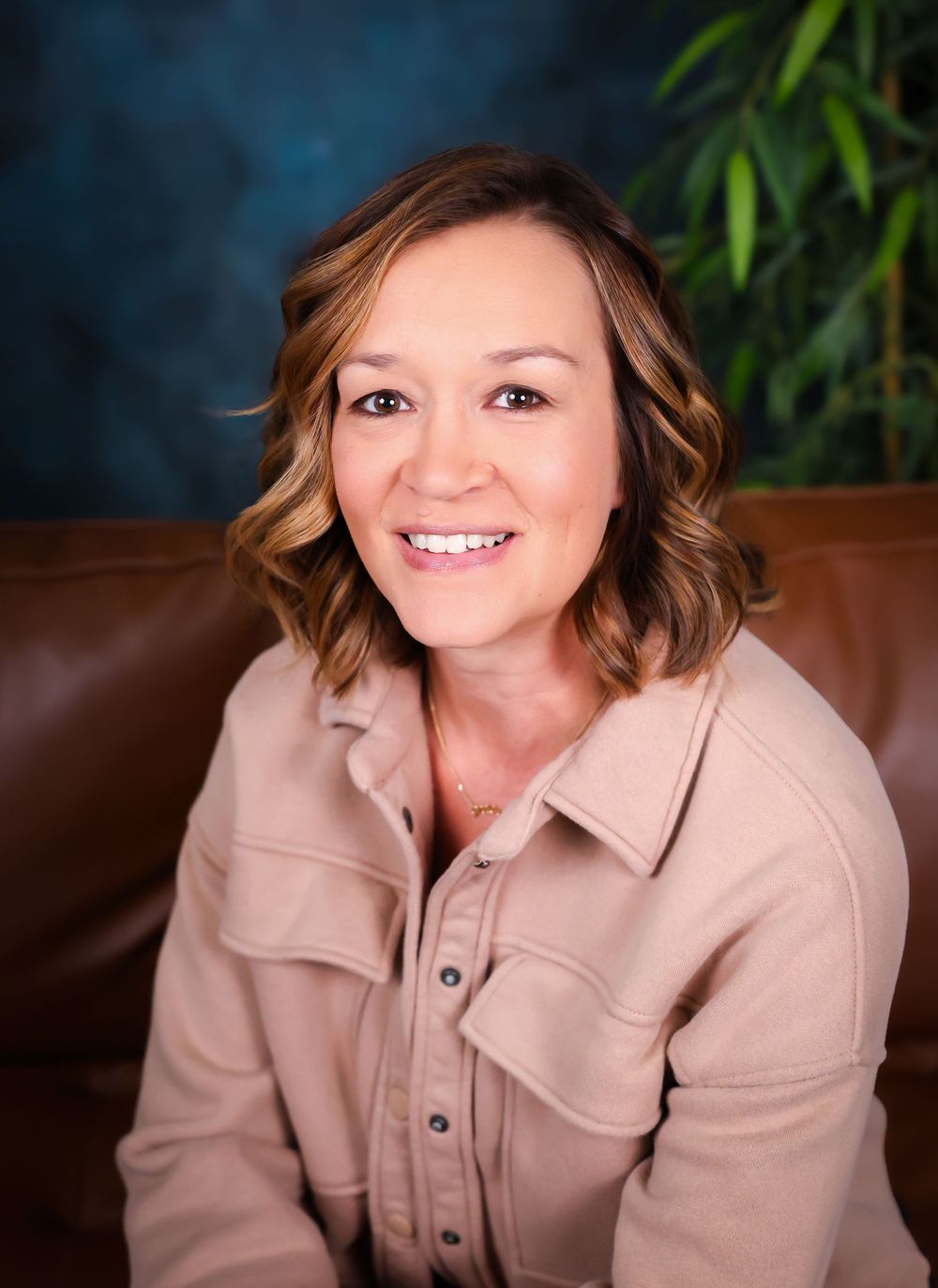 Professional headshot with beige jacket against natural background.