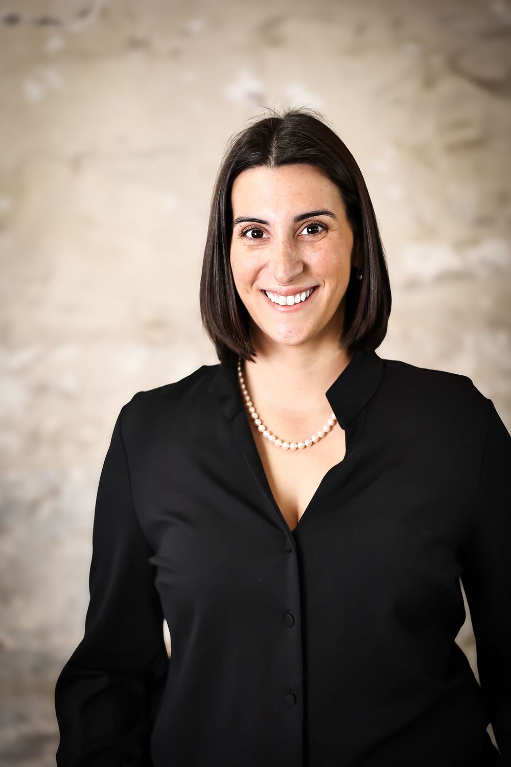 Corporate headshots in black blazer with pearl necklace against neutral background.