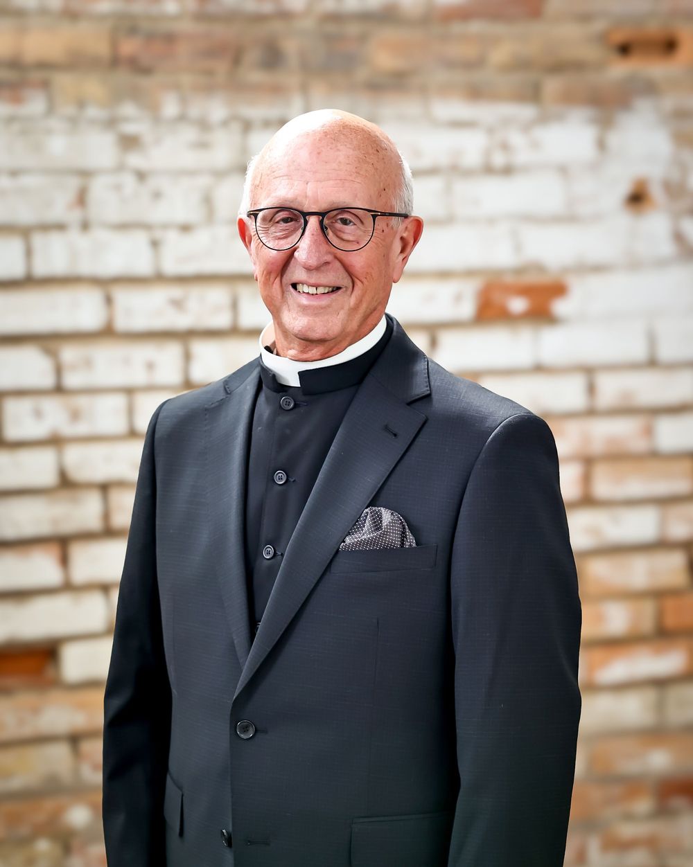Formal business portraits against brick wall wearing classic black suit and glasses.