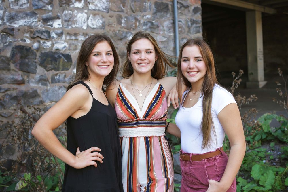 Three young women pose together outdoors in summer attire against a stone wall background.