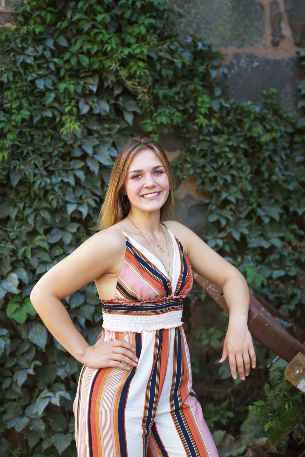 A person in a striped summer dress poses against an ivy-covered wall outdoors.