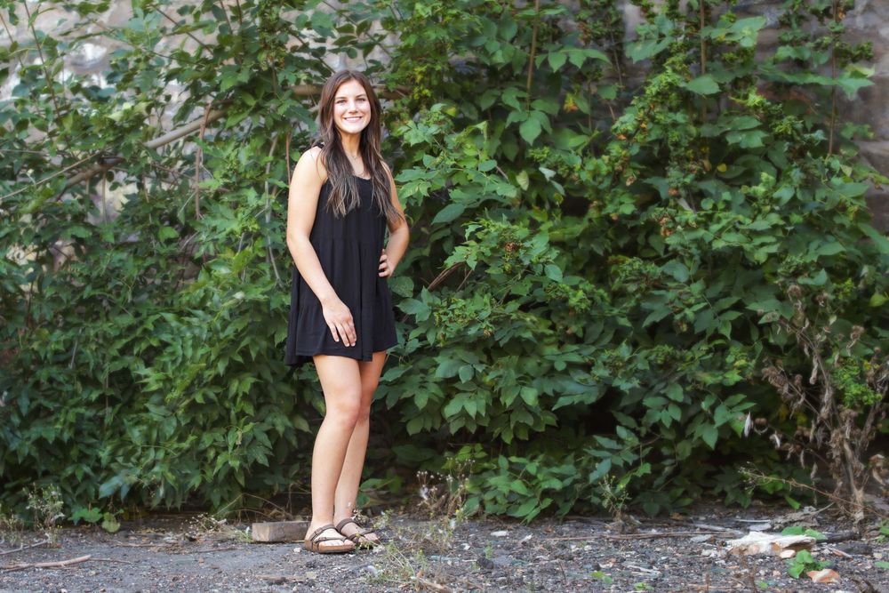 A person in black clothing poses outdoors near green foliage and trees in a natural setting.