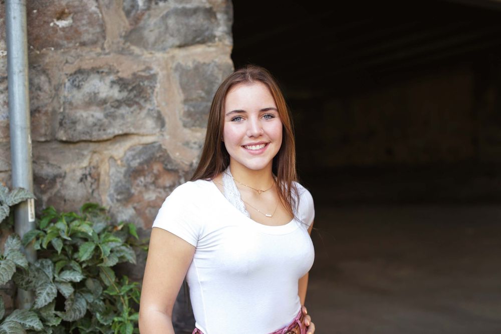 A person in a white top smiles while standing against a rustic stone wall with climbing ivy.