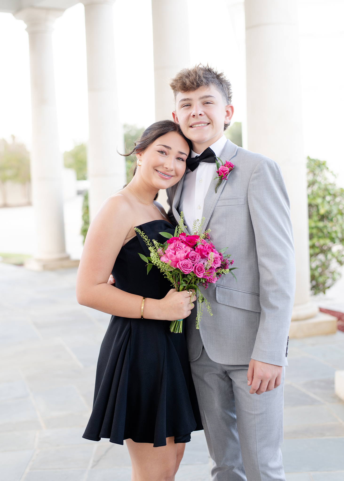 Teenage couple dressed for a formal dance, girl in black dress holding pink roses, boy in gray suit.