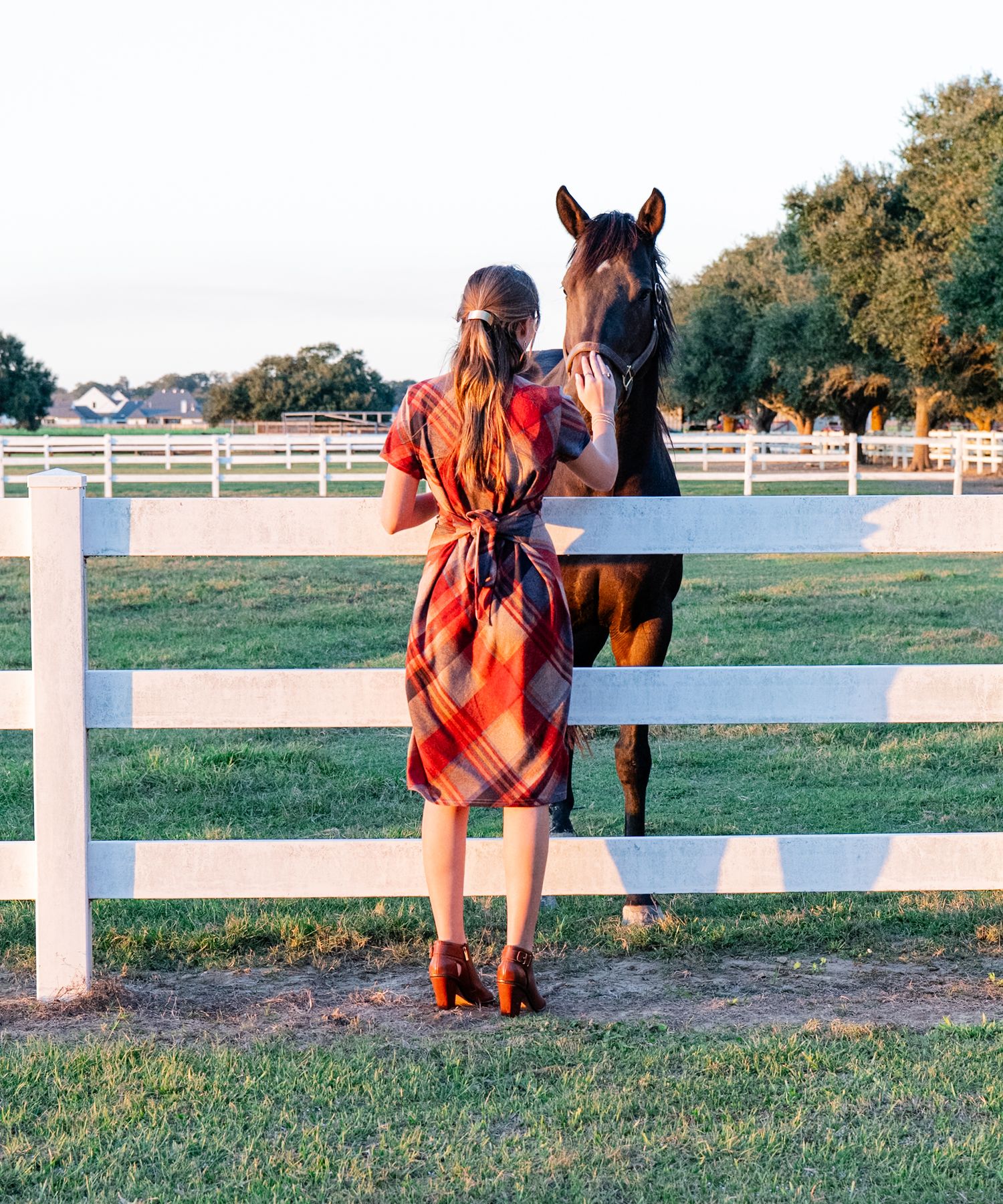 Woman in a red plaid dress standing at a white fence petting a brown horse at a sunny farm.