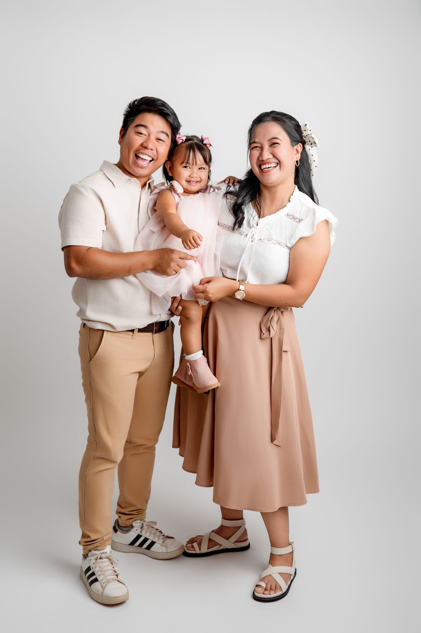 A family poses together in coordinated beige and blush outfits against a white studio backdrop for a portrait session.