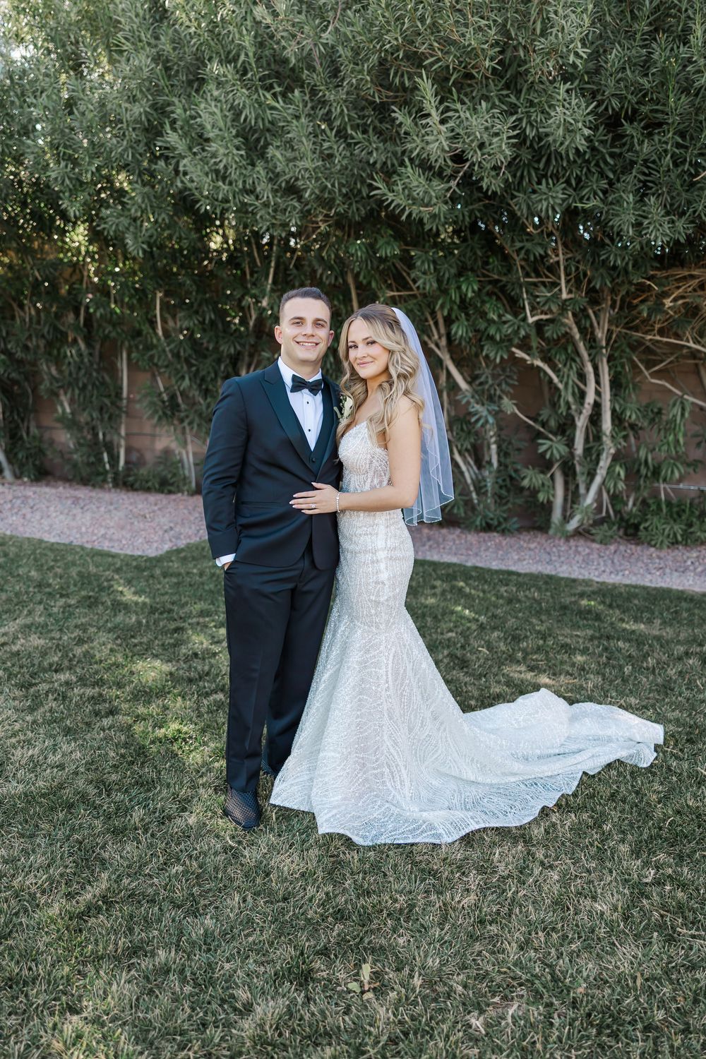 A wedding couple poses together on a lawn with greenery and trees in the background.