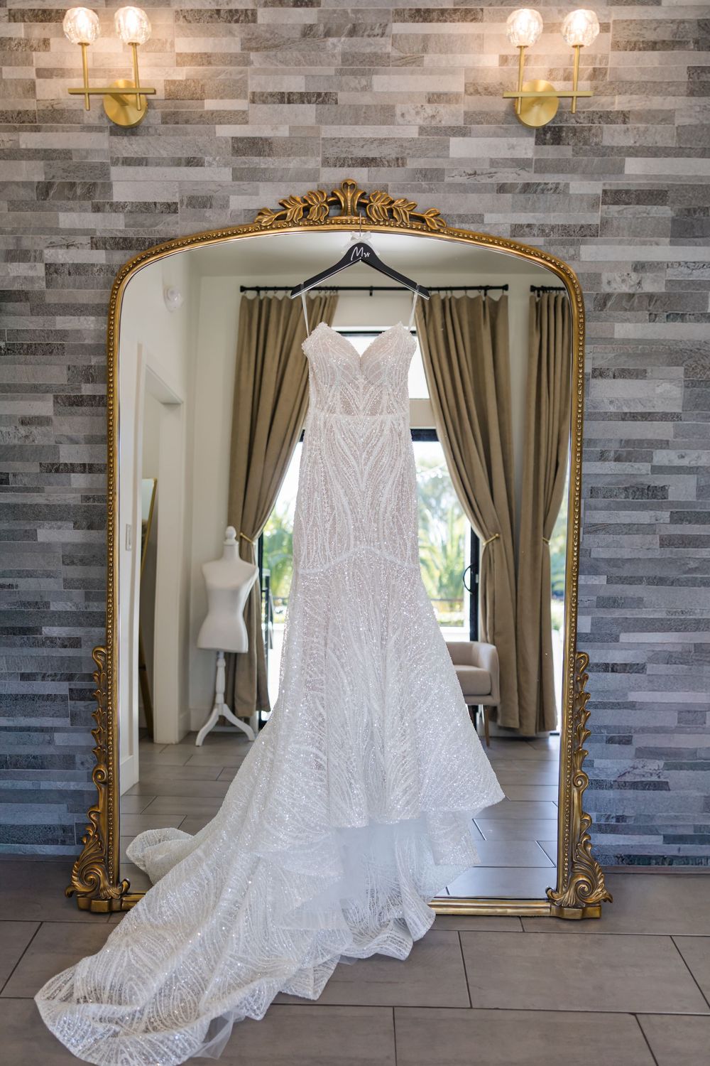 White lace wedding dress displayed in front of elegant gold-framed mirror against grey stone wall with sconce lighting.