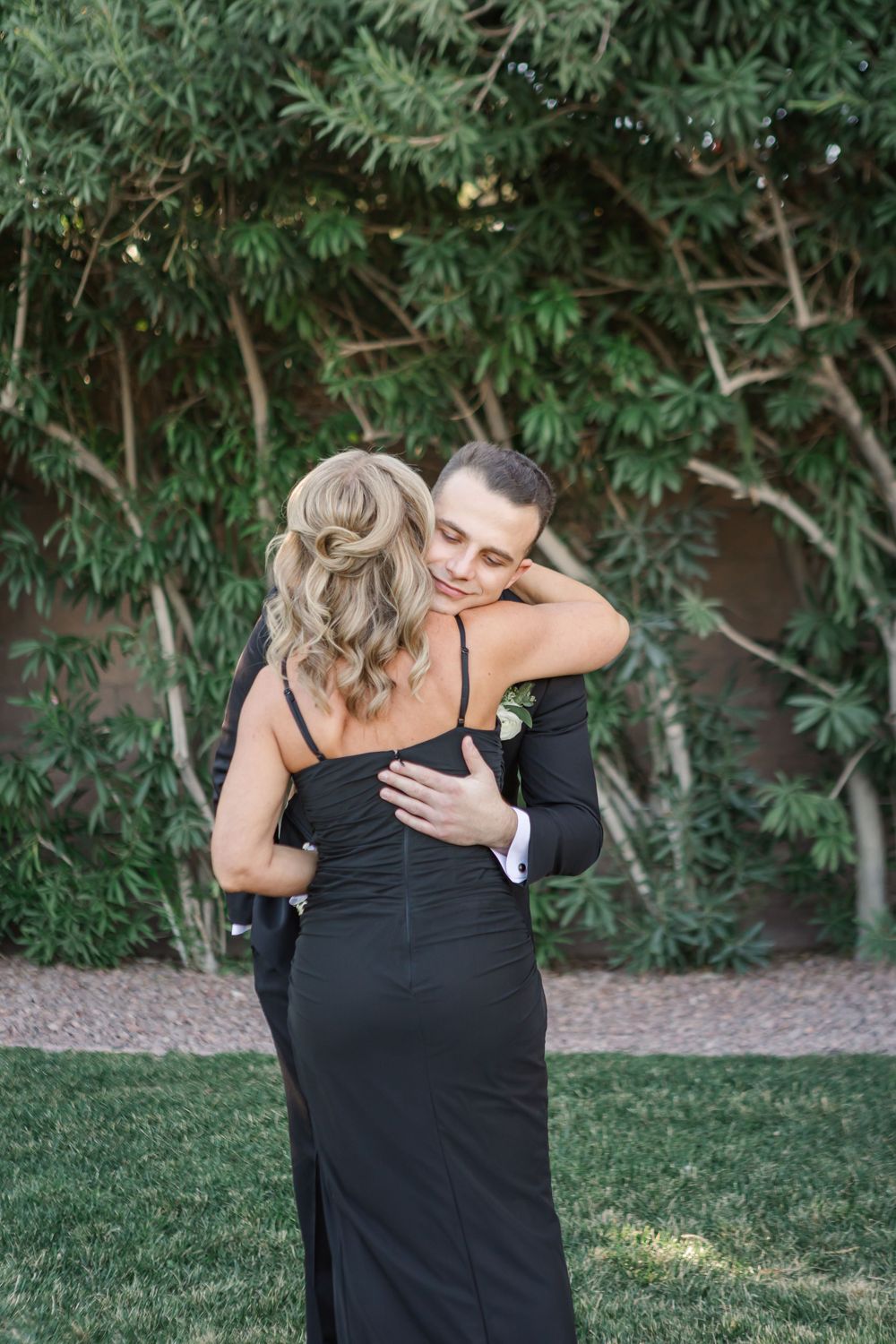 A couple shares an emotional embrace outdoors in a garden setting with green foliage in the background.