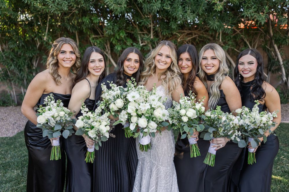A wedding party in black dresses poses together with white floral bouquets in an outdoor garden setting.