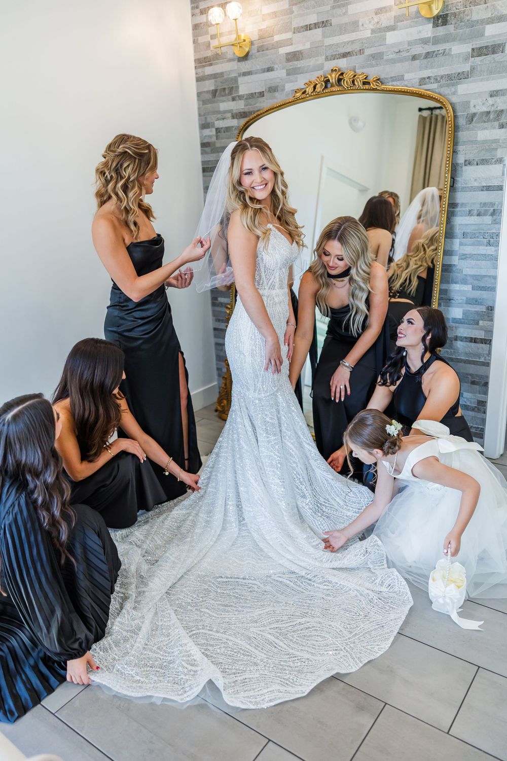 Bridesmaids in black dresses help arrange the train of an intricate lace wedding gown during pre-ceremony preparations.