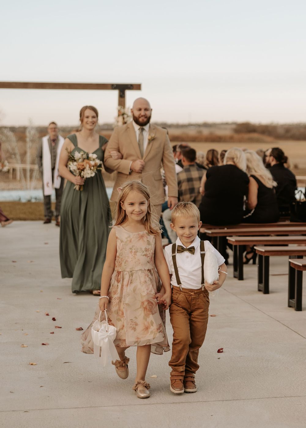 Two young children walk hand in hand on a paved path at an outdoor venue with wedding guests in sage green dresses nearby.