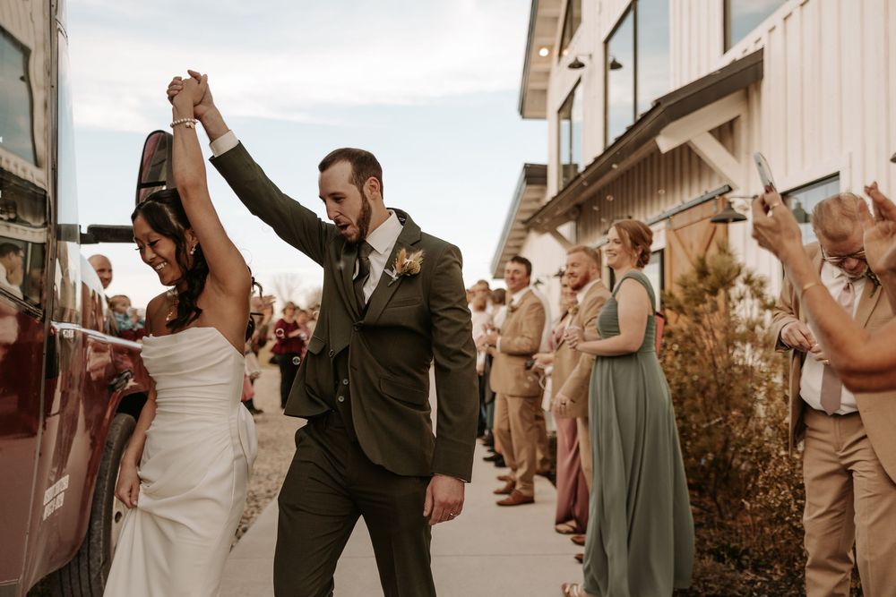 Wedding guests raise their arms in celebration as newlyweds walk down a rustic outdoor path at sunset.
