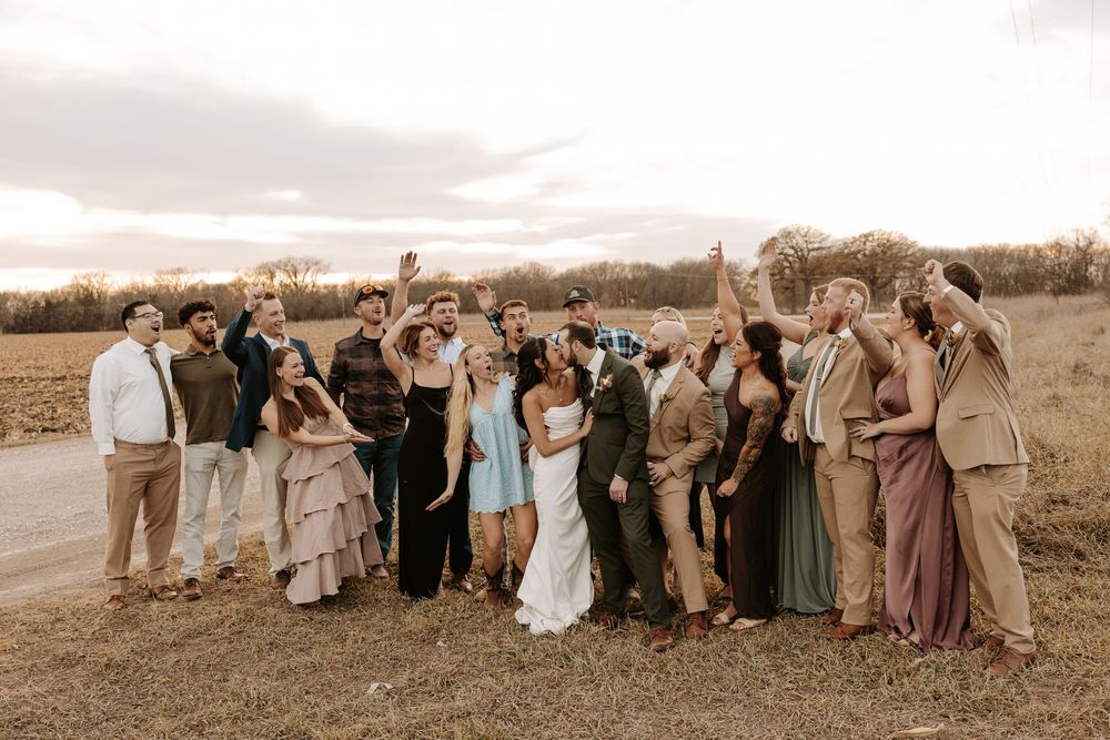 Wedding guests gather for a group photo in a scenic outdoor field at sunset.