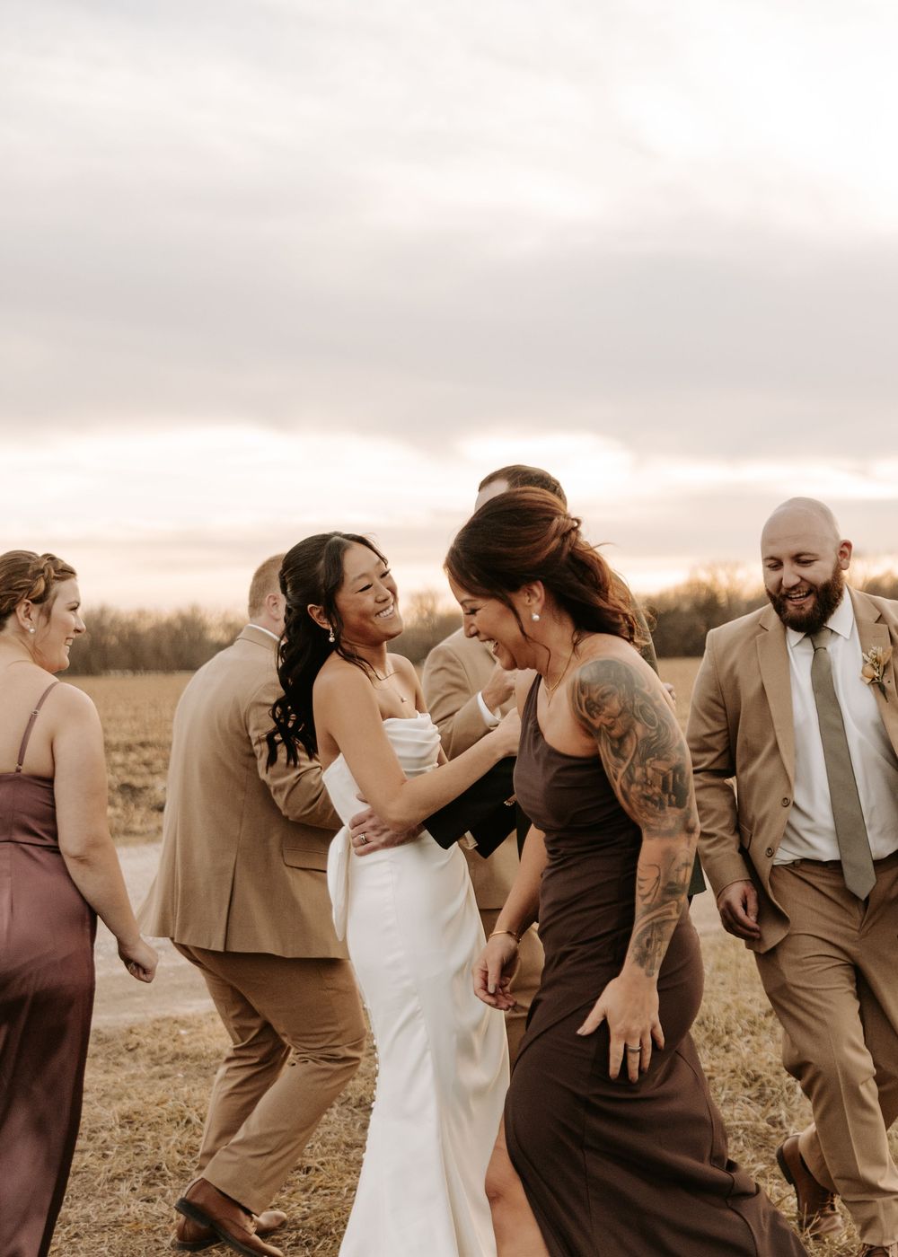 Wedding party dancing and laughing together in a rustic outdoor field at sunset.