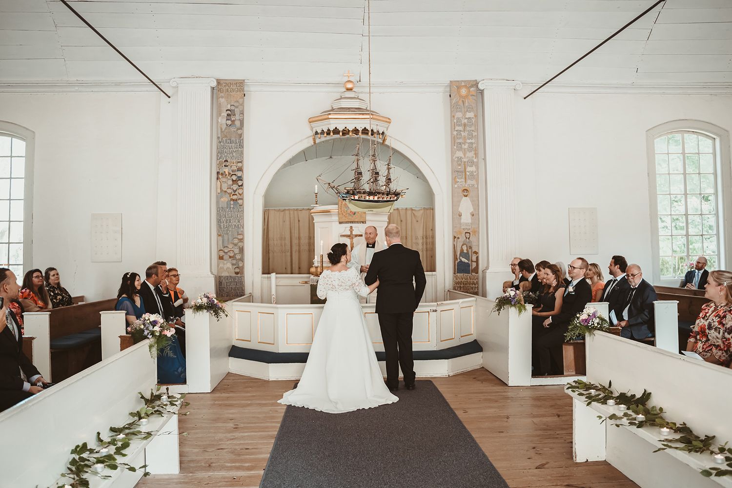 Bröllop Medevi brunnskyrka, brudpar står framför prästen i den vita gamla kyrkan, gästerna sitter i kyrkbänkarna. Bröllopsfoto Lets Bloom