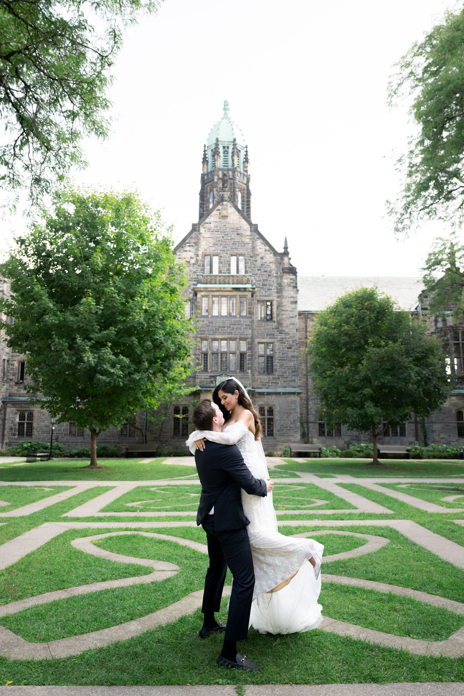 A couple embraces on a manicured lawn with gothic university buildings in the background.