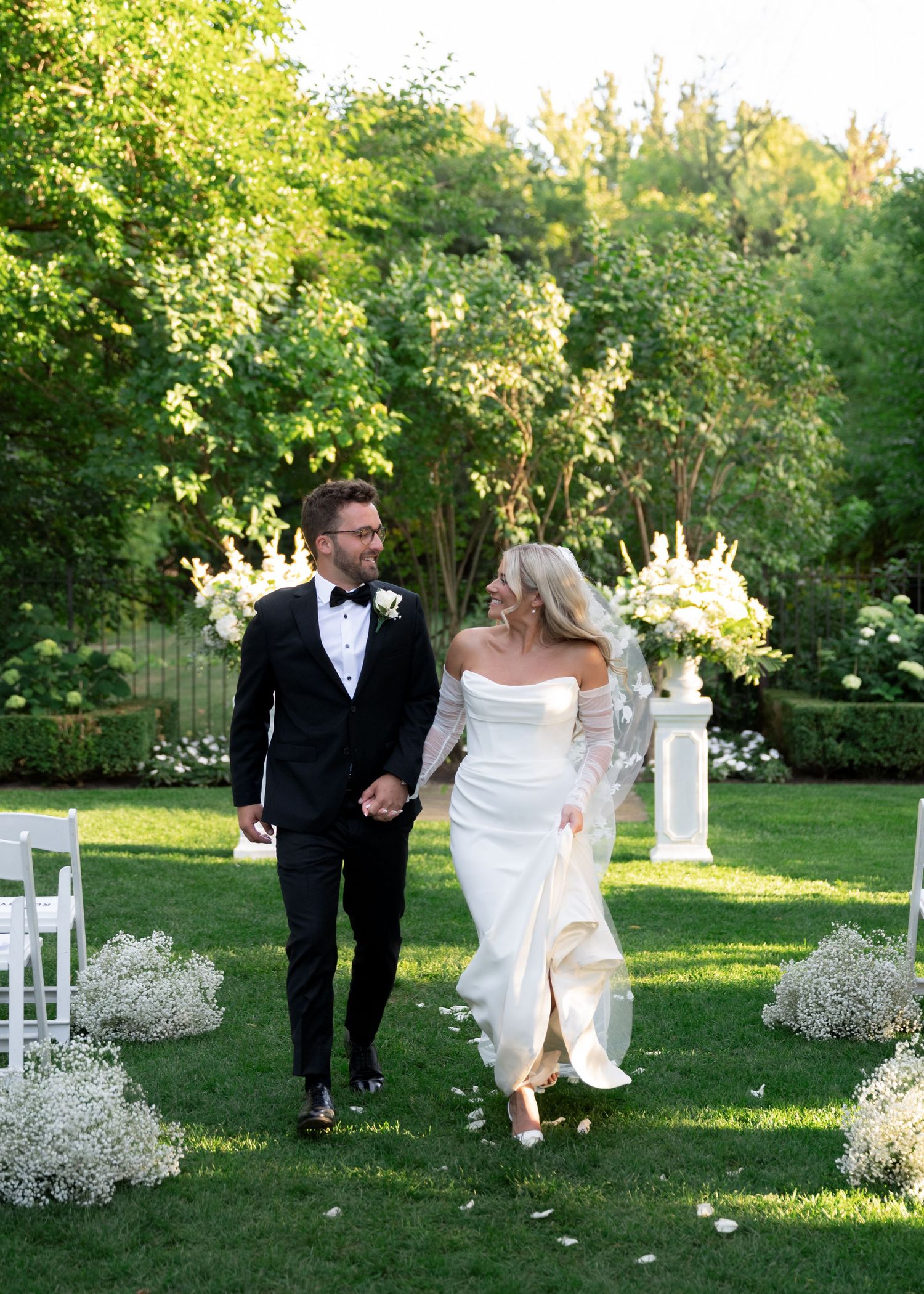A couple in formal wedding attire joyfully walks across a sunlit garden lawn lined with white chairs and baby's breath flowers.