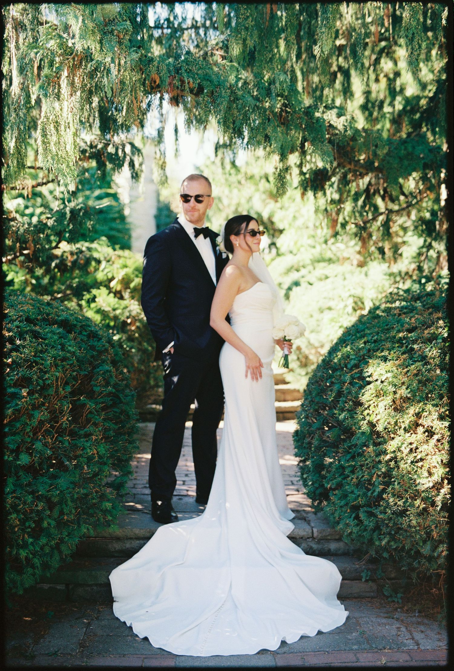 A wedding couple poses on a garden path beneath Spanish moss and greenery in an elegant outdoor setting.