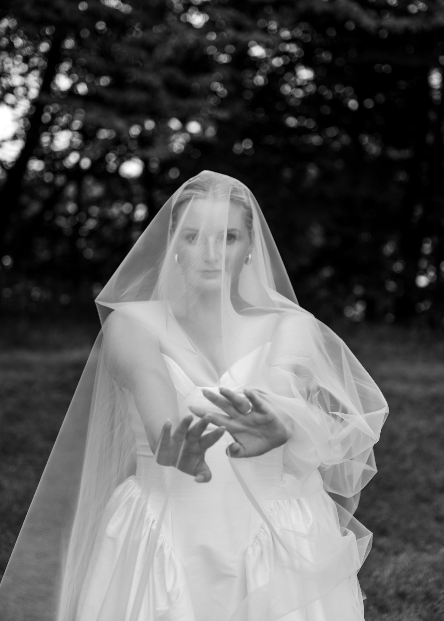 Black and white photo of a bride in a white gown and veil, holding her hands forward in a misty outdoor setting.