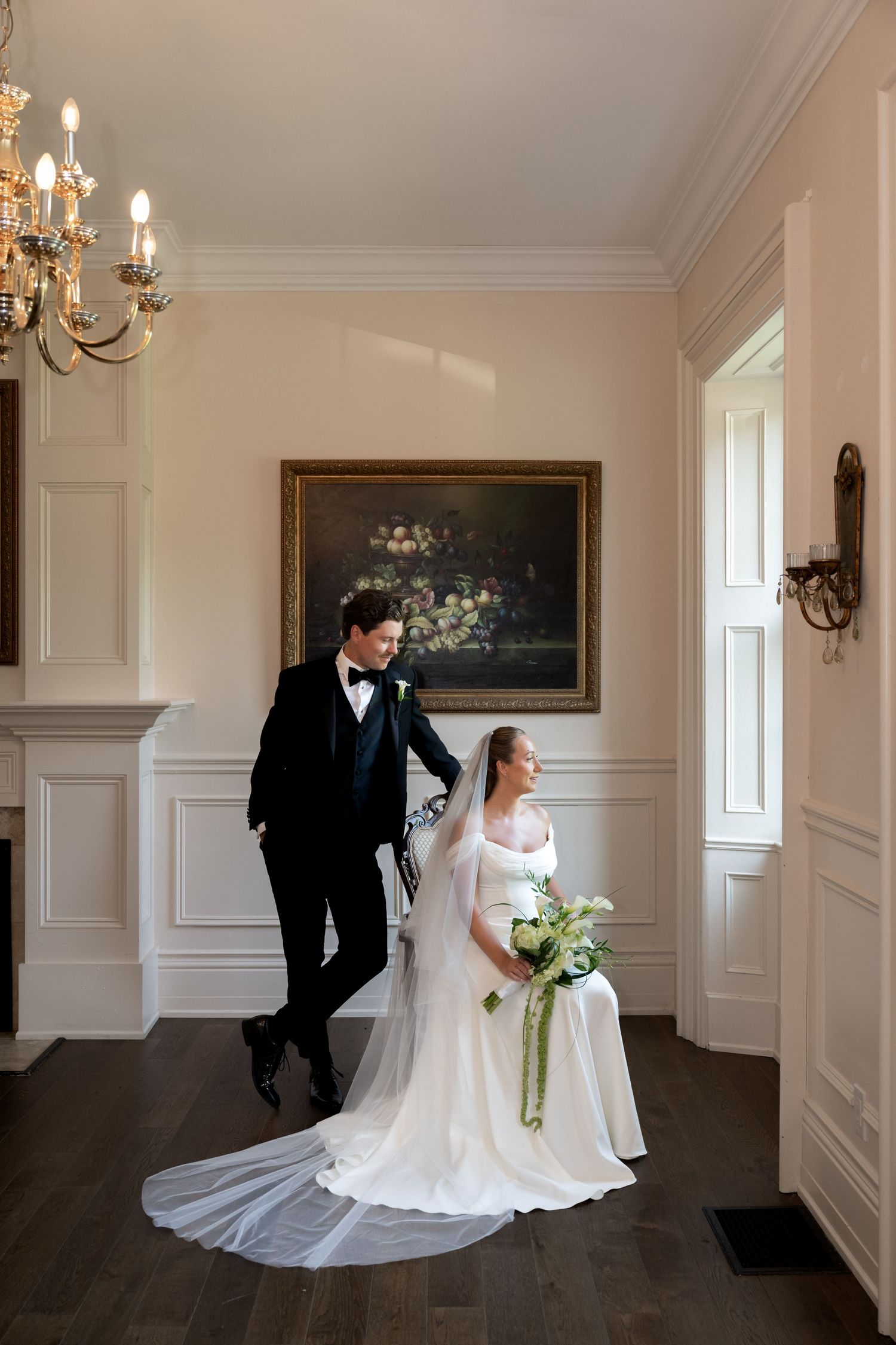 Elegant bride and groom pose in a classic white-paneled room with chandelier, bride holding a white bouquet with green ribbon.