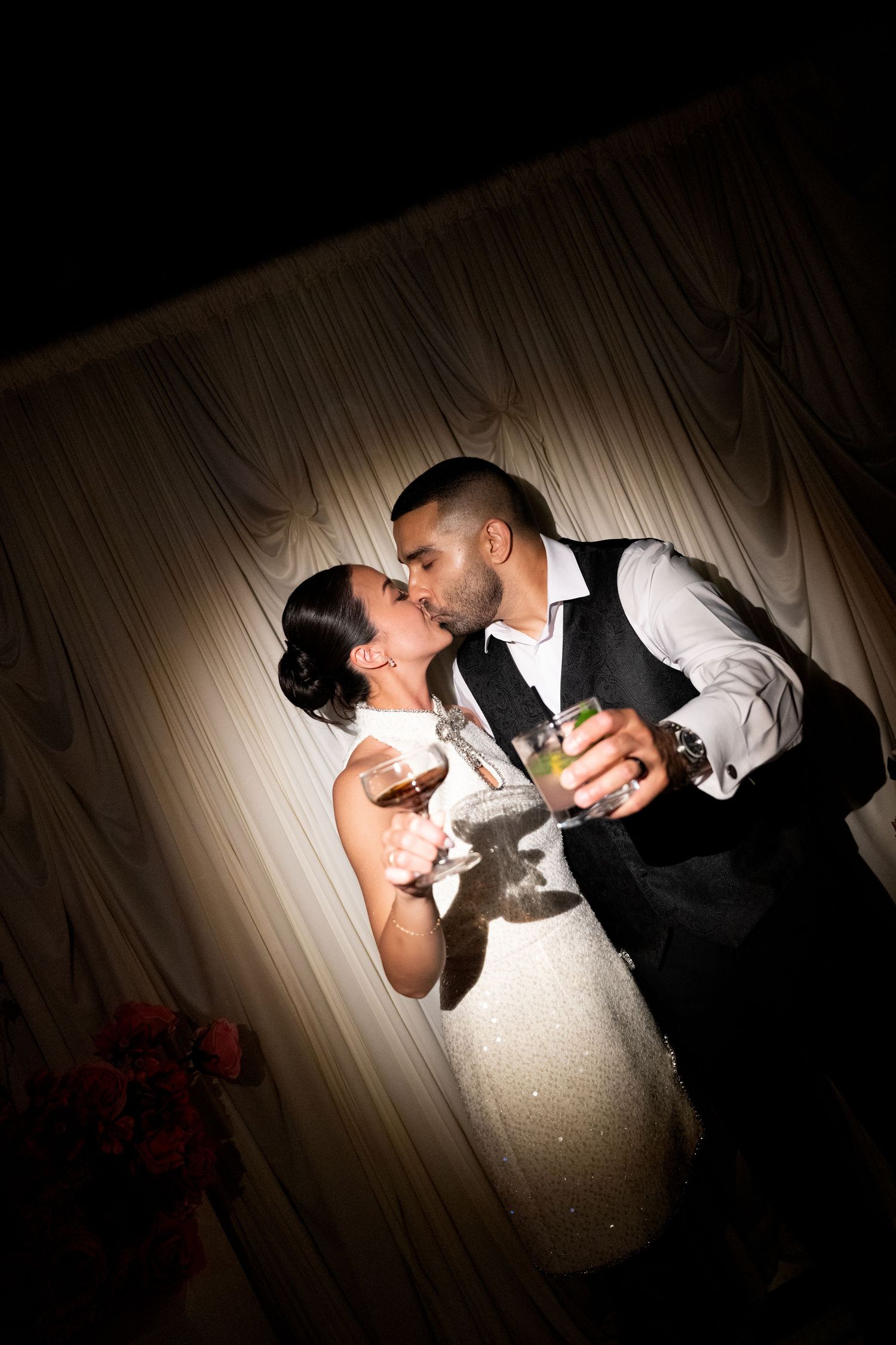 A newlywed couple shares a romantic kiss while holding champagne glasses at their wedding reception.