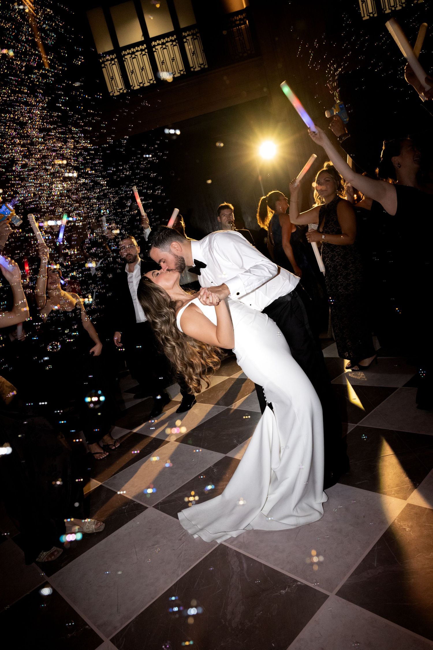 Bride and groom share a romantic dip kiss on a checkered dance floor surrounded by guests holding glowing light sticks.