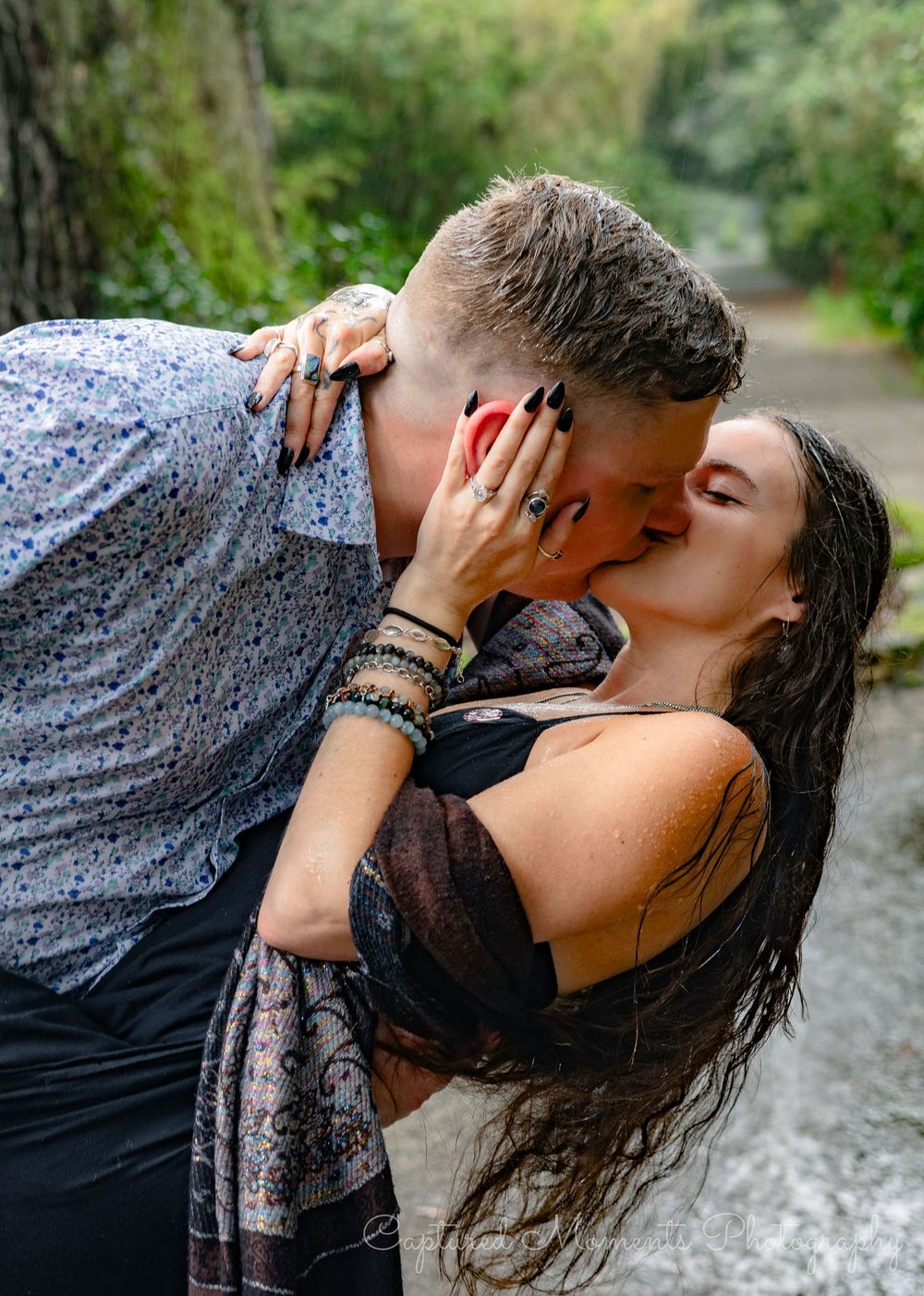 A romantic couple shares a passionate kiss on a nature path surrounded by lush greenery.