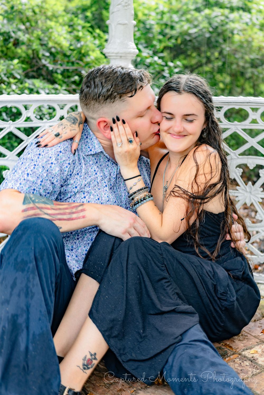 A couple shares tender kisses while sitting on a white lattice bench in a lush garden setting.