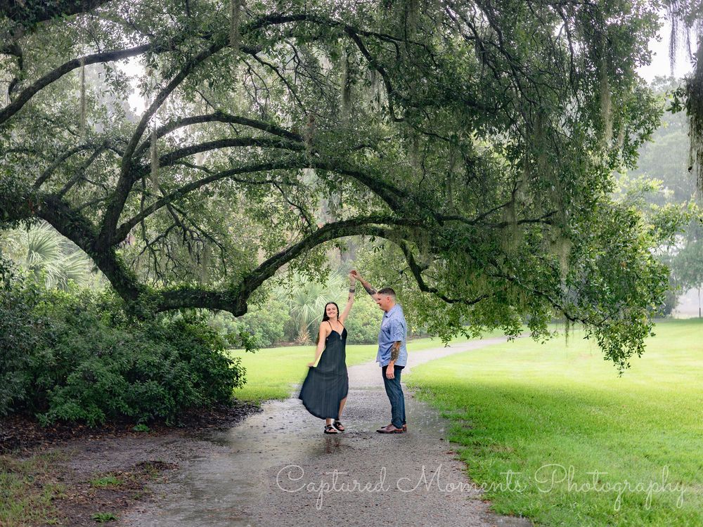 Two people stand together beneath a large drooping willow tree on a path in a lush green park setting.