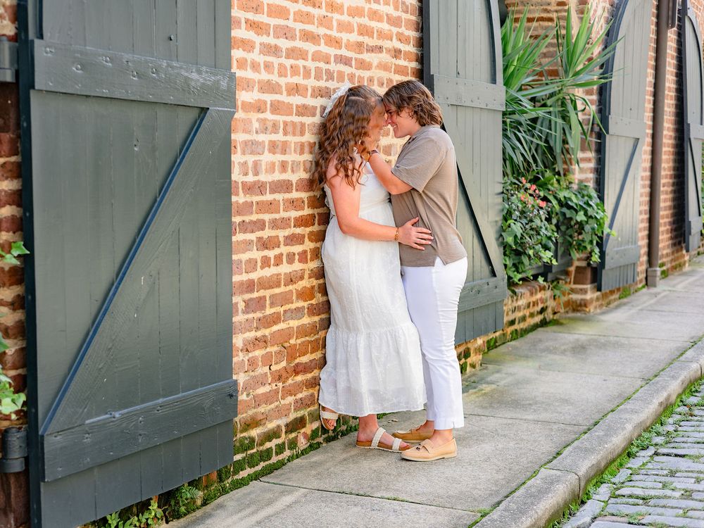 A couple in white outfits share a tender moment along a brick wall pathway.