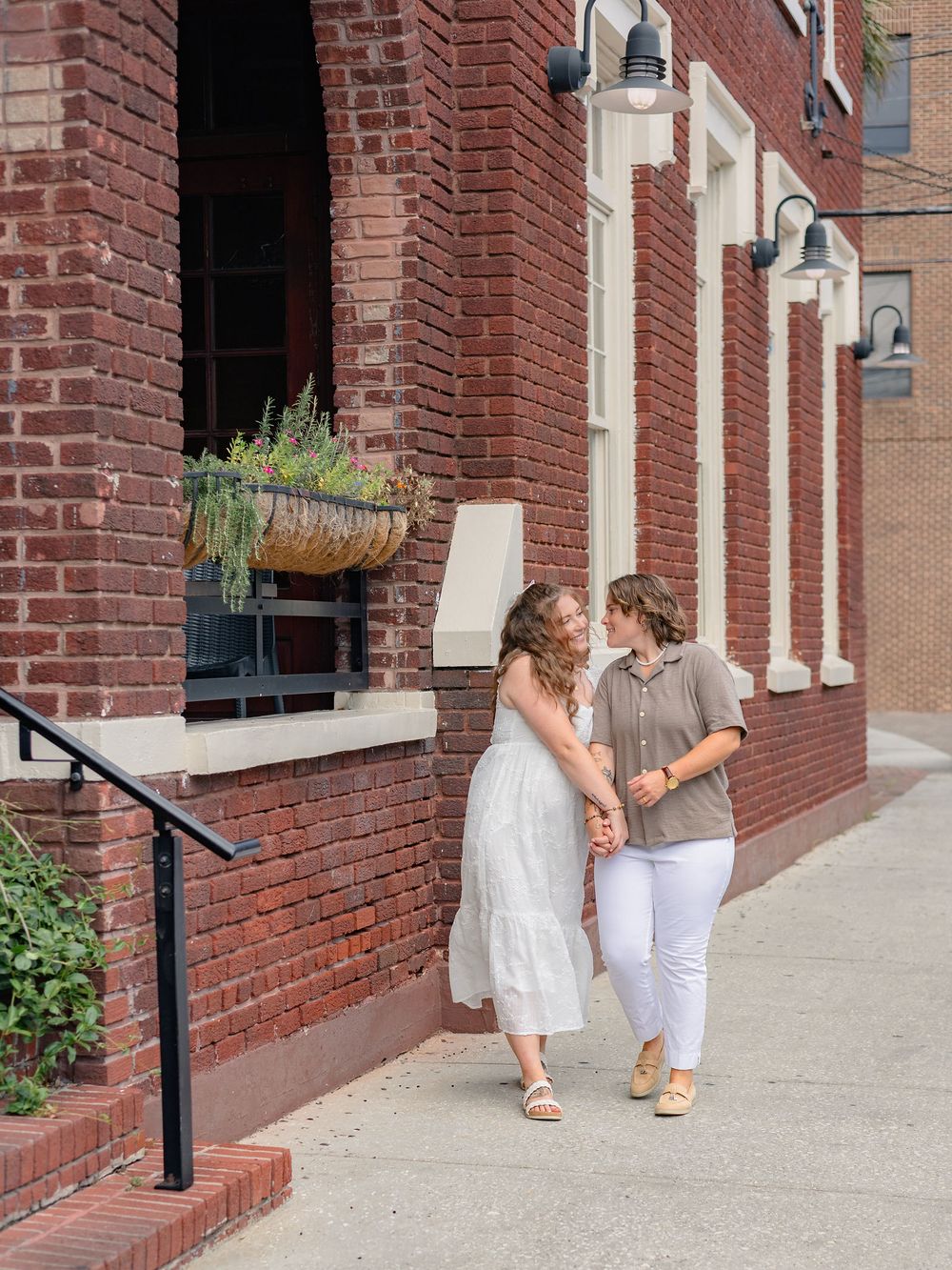 Two people in light summer outfits walking together along a brick building with hanging planters and white columns.