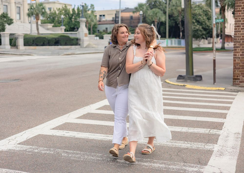Two people walk arm in arm across a crosswalk on a sunny day, one wearing white and one in casual attire.