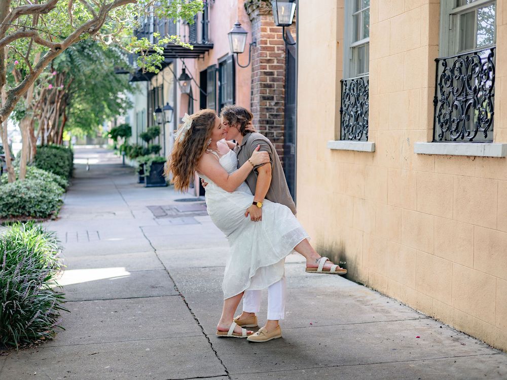 A couple embraces and kisses on a charming city sidewalk with peach-colored walls and lush greenery.