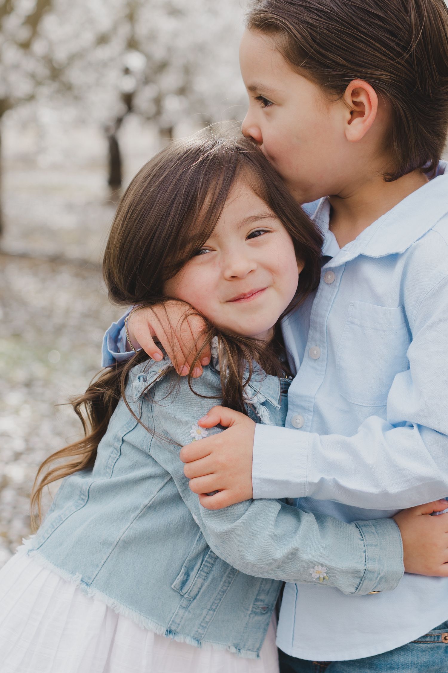 Two children in light blue clothing share a sweet embrace against a light colored background.