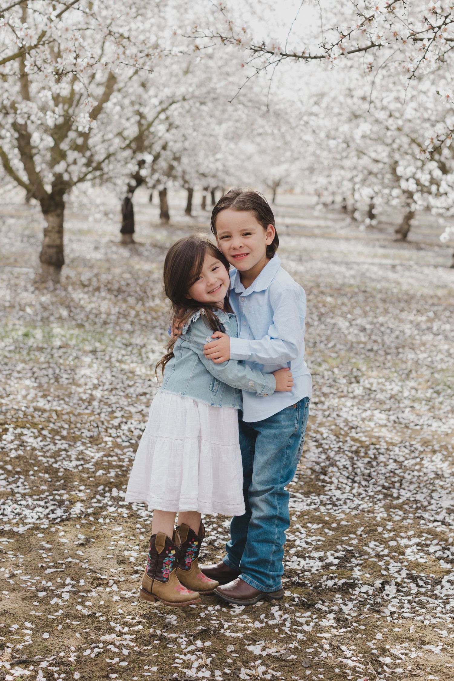 Two children in light blue and denim share sweet embraces while playing among fallen white flower petals in an orchard.