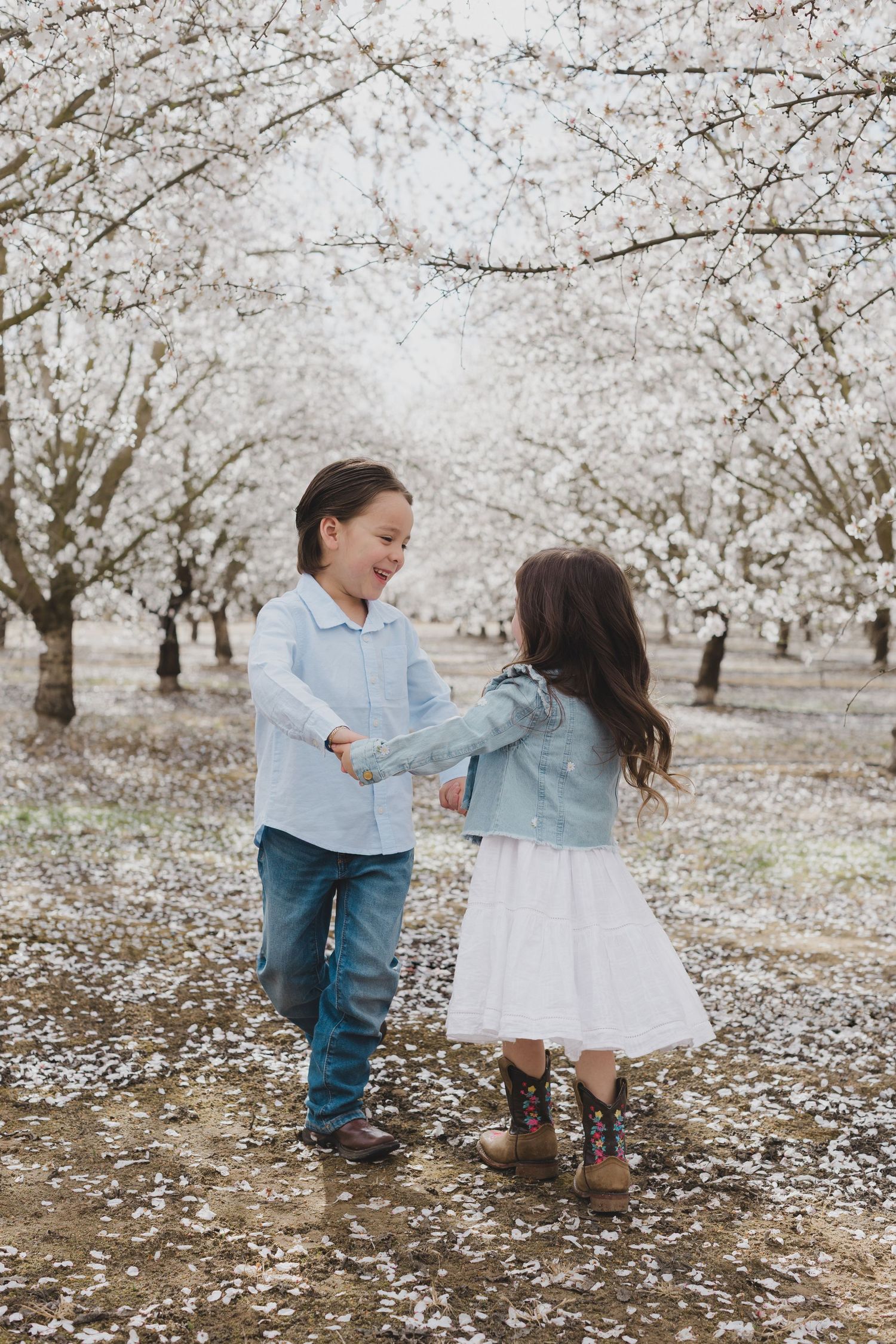Two children dance together in an orchard of blooming cherry trees with white petals scattered on the ground.
